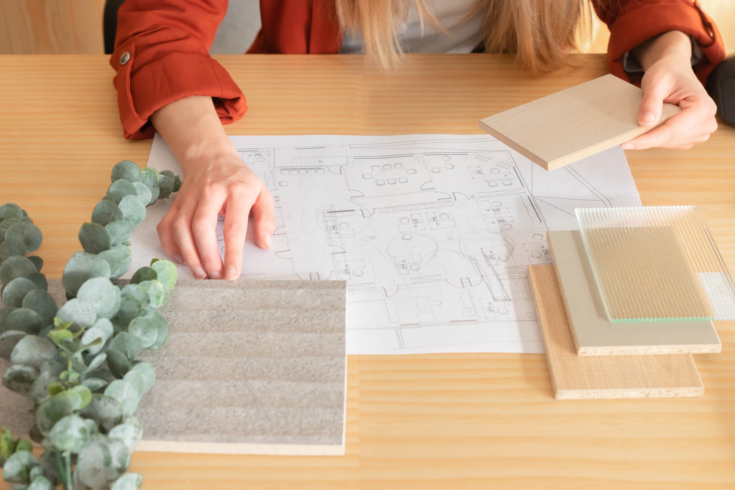 Innenarchitektur mit Materialmustern und Pflanze auf einem Tisch, während eine Person mit einem Holzmuster arbeitet.